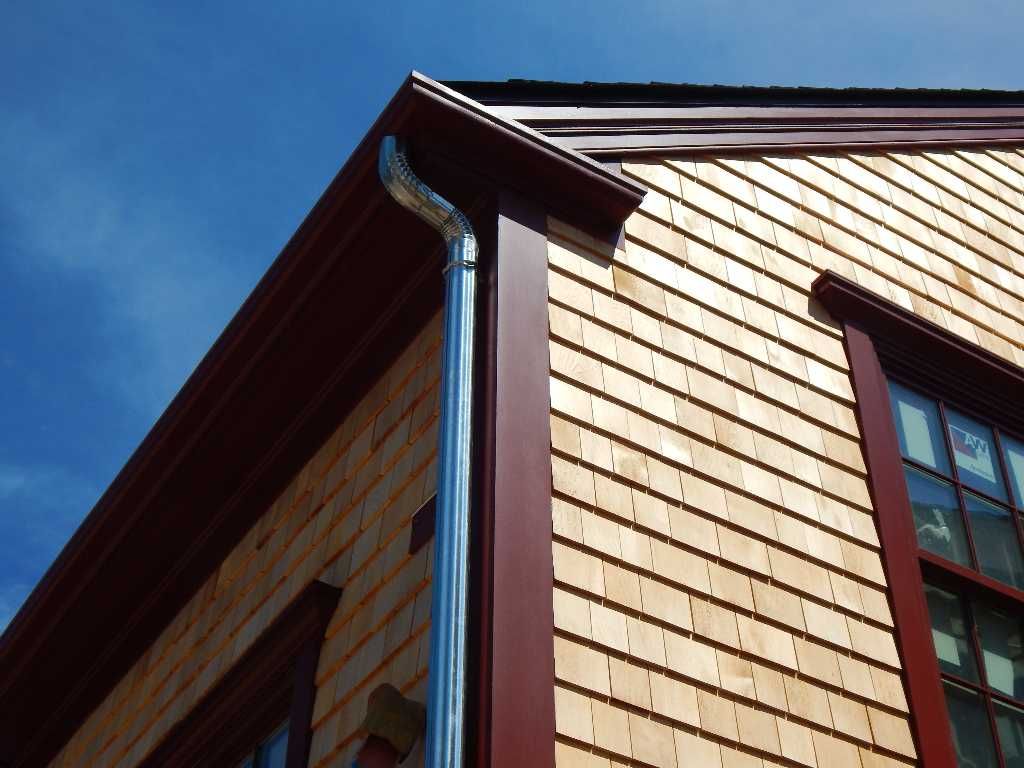 Exterior corner of a building with cedar shake siding, dark red trim, and a metal downspout against a blue sky.