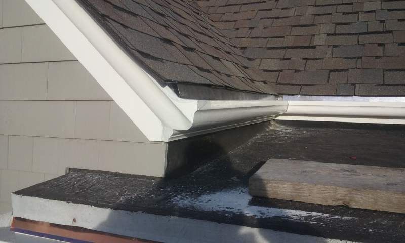 Close-up of a house roof corner with white gutters, asphalt shingles, and gray siding.
