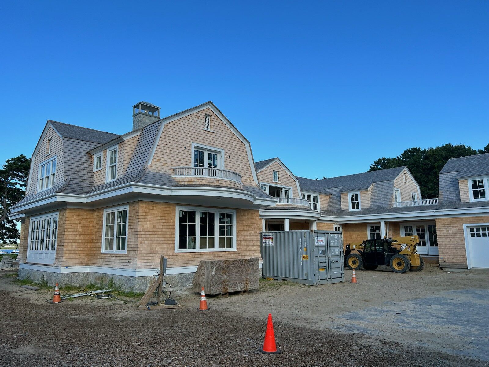House under construction with cedar shingle roof and siding, on a gravel lot, under a blue sky.