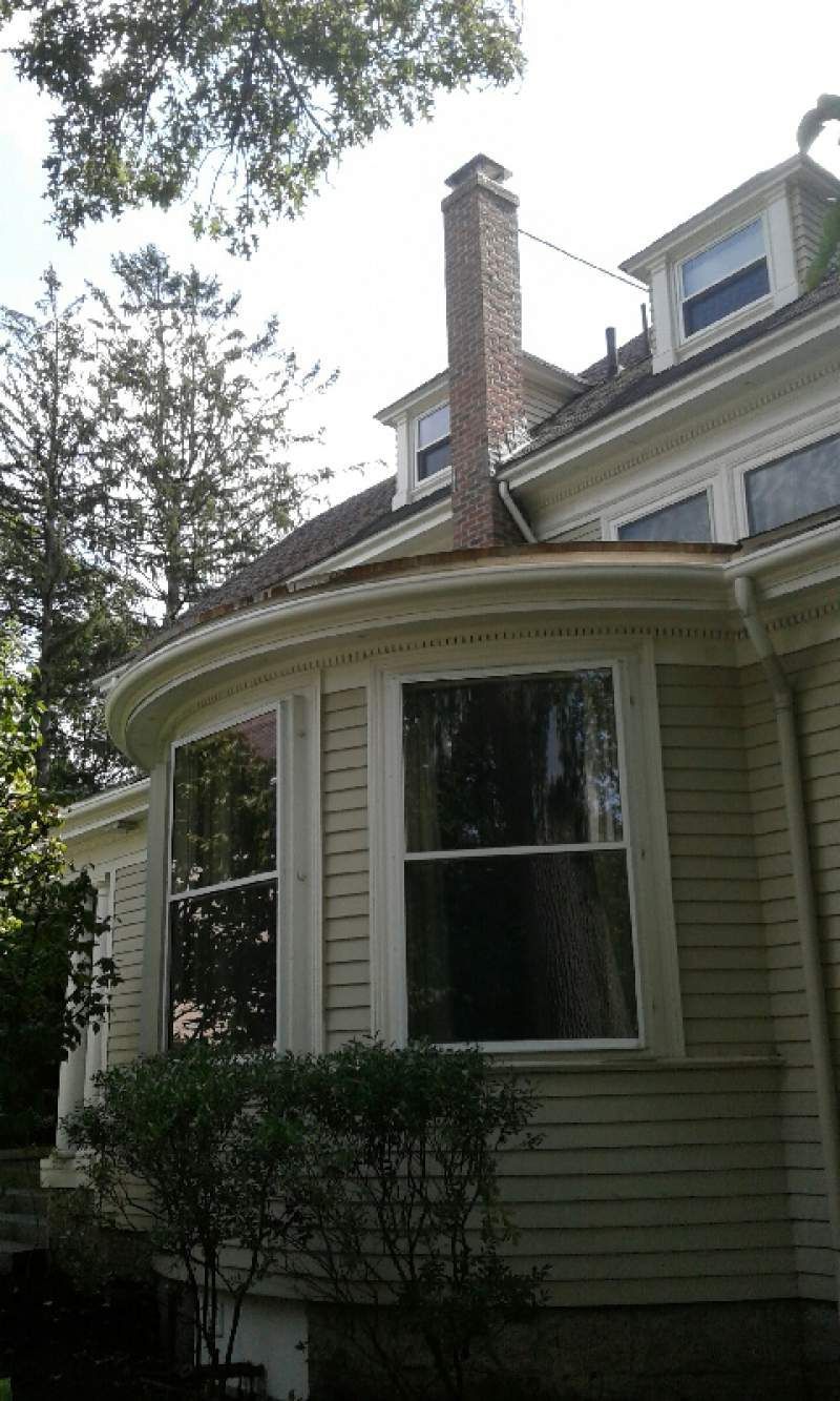 Cream-colored house with curved bay windows and a brick chimney on the roof, surrounded by trees.