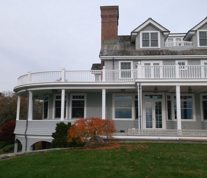 Gray house with white trim, balcony, and chimney on a grassy hill; a tree with red leaves is in front.
