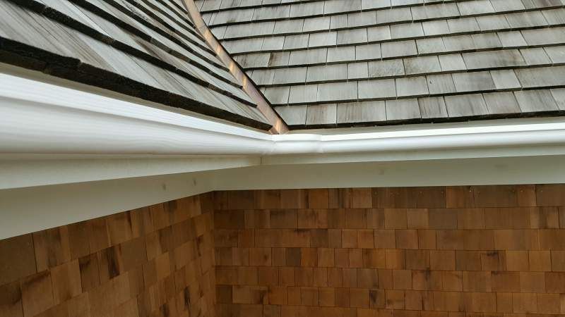 Brown wooden shingle roof with white trim and gutter against a brown, textured exterior wall.