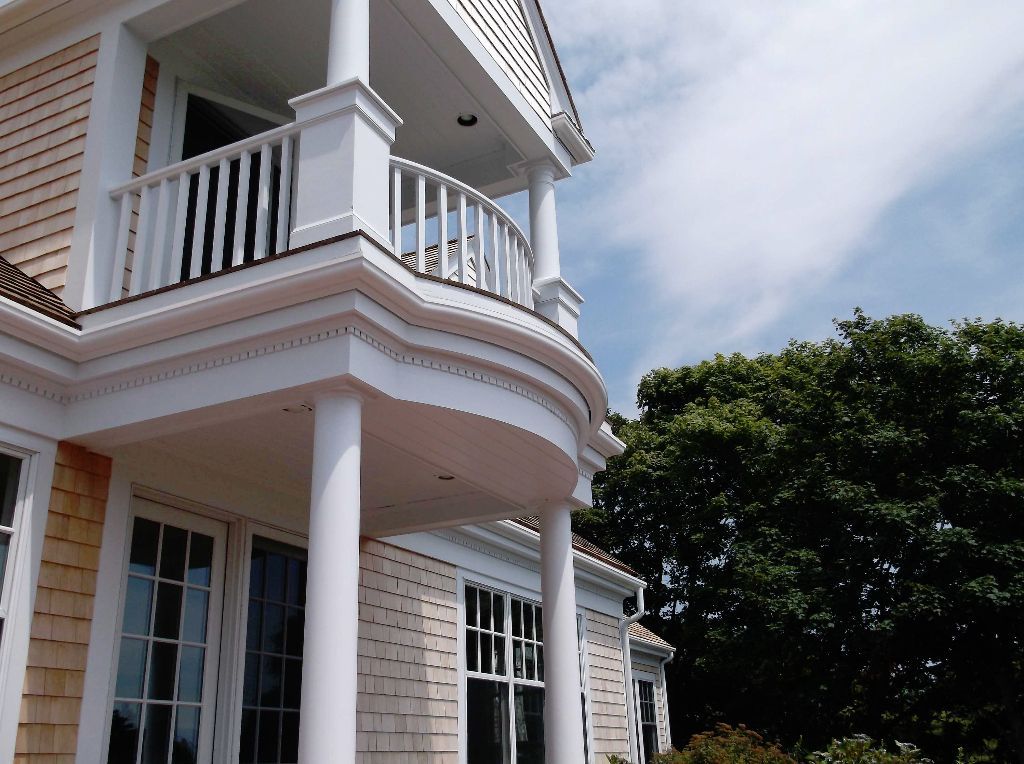 White balcony with curved railing and columns on a beige shingled house, blue sky and trees.