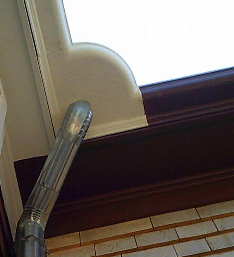 Metal downspout draining rainwater from a white gutter, adjacent to a window.