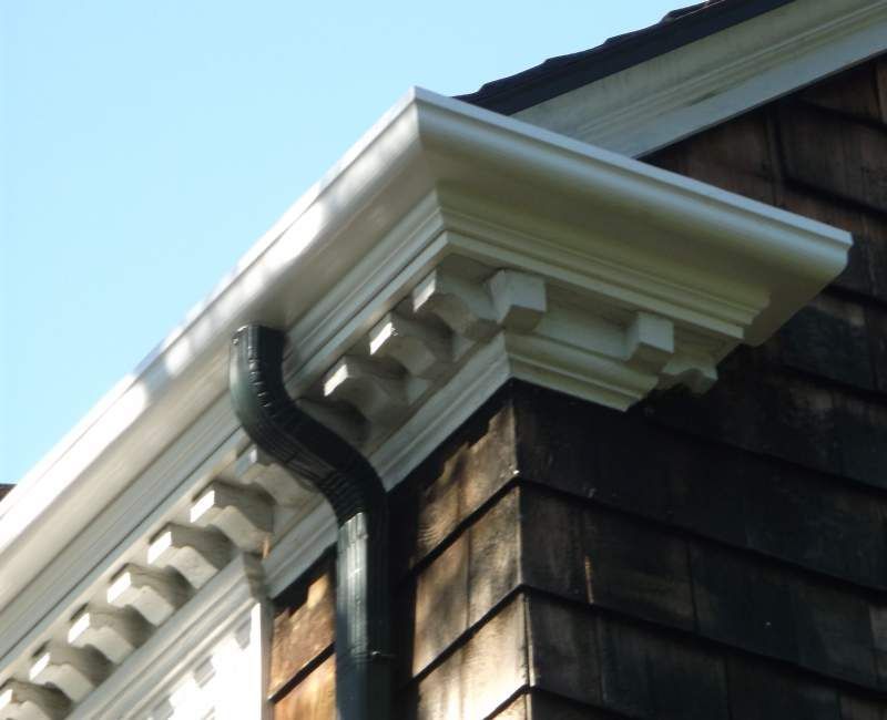 Corner of a house with white trim, dark shingles, and a black downspout against a blue sky.