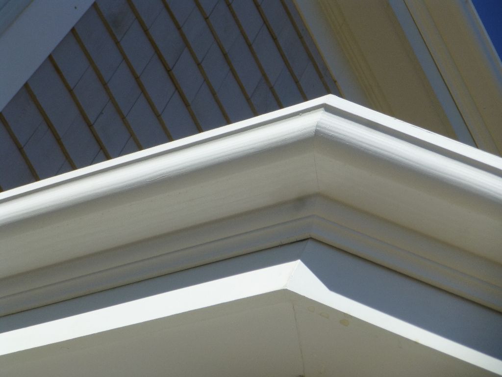 White corner of a house, showing a gutter and siding under a blue sky.