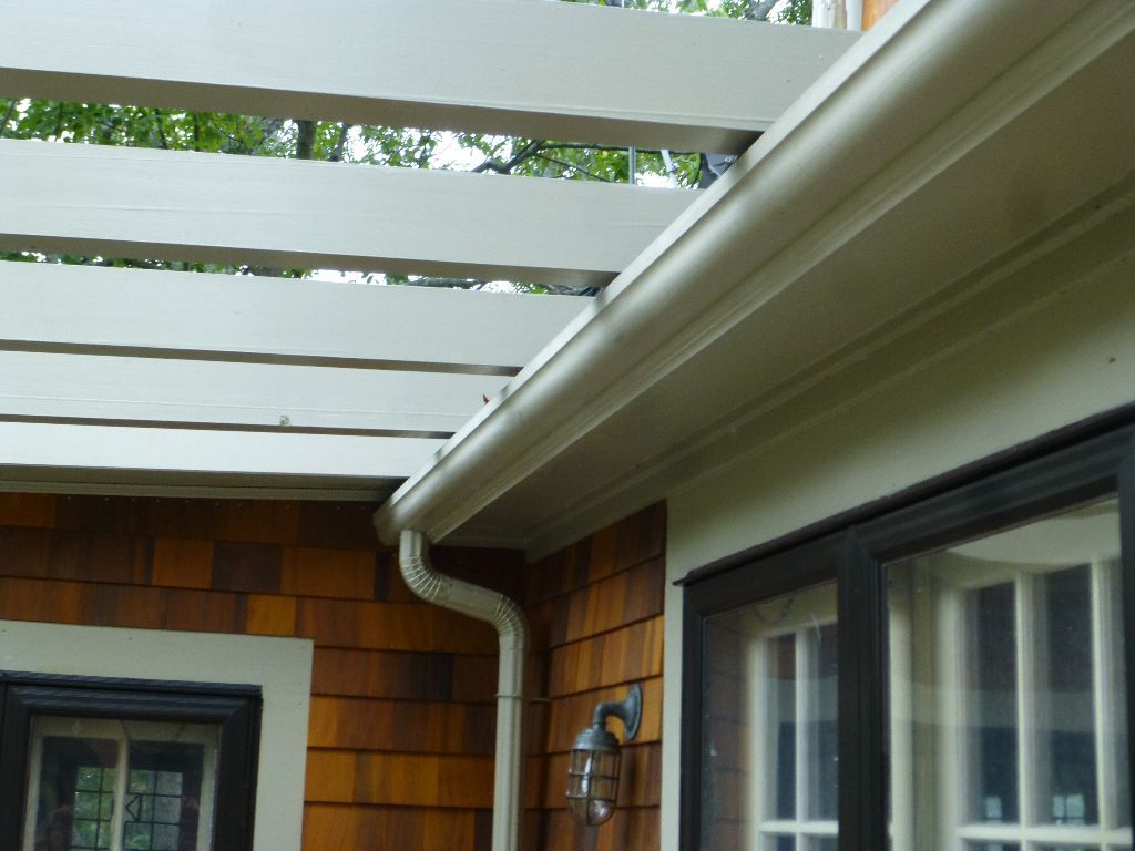 Guttering on a house with a pergola above, brown shingles and windows with black frames.
