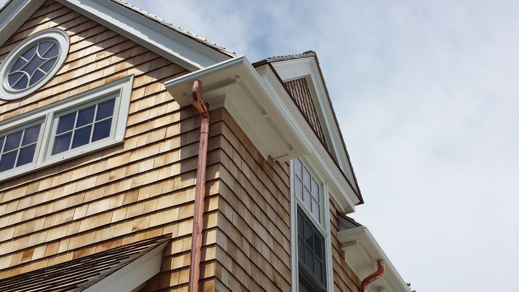 Cedar shingle siding on a house corner with copper gutters and white trim under a cloudy sky.
