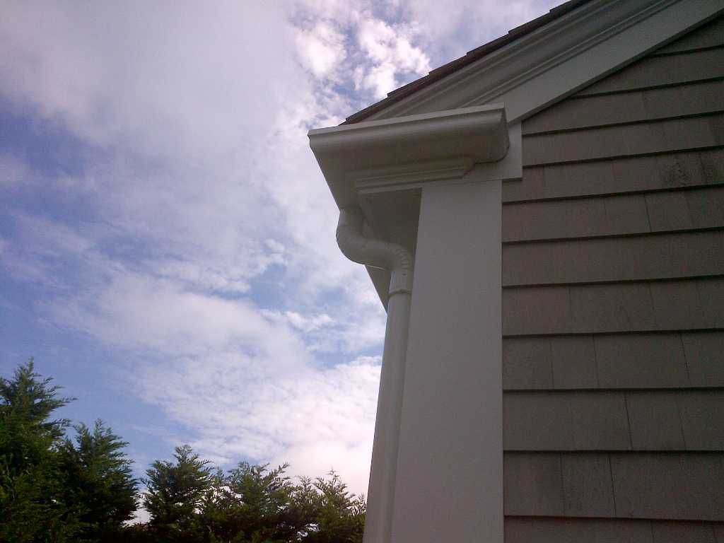 White gutter and downspout on a building with gray shingle siding, under a partly cloudy sky.
