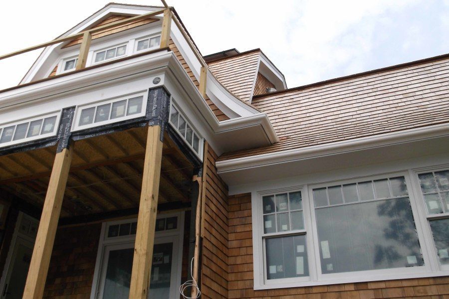 Exterior view of a house under construction with brown cedar shake siding, white trim, and a partially built porch.