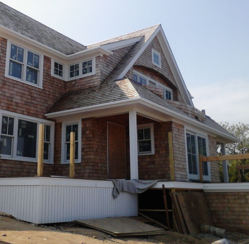 Exterior of a house under construction; cedar shake siding, white trim, multiple windows, porch with wood supports.