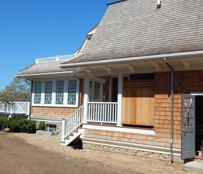 A two-story shingle-sided house with a porch, stairs, and a wooden door on a sunny day.