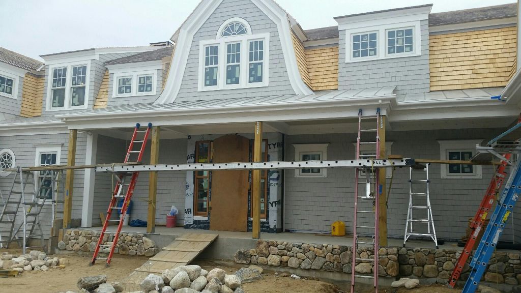 House under construction; gray siding, tan roof, ladders, scaffolding, and stone foundation.