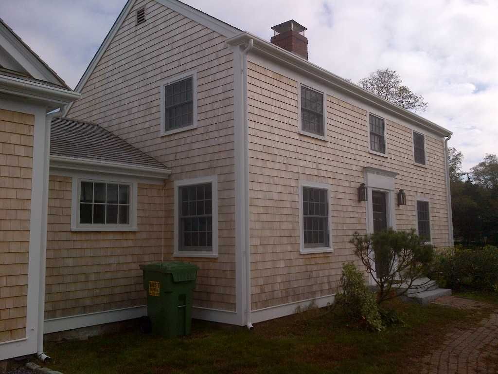 Two-story wood-shingled house with white trim, windows, and a green trash can outside.
