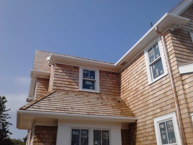 Two-story house with cedar shake siding, white trim, and a clear blue sky background.