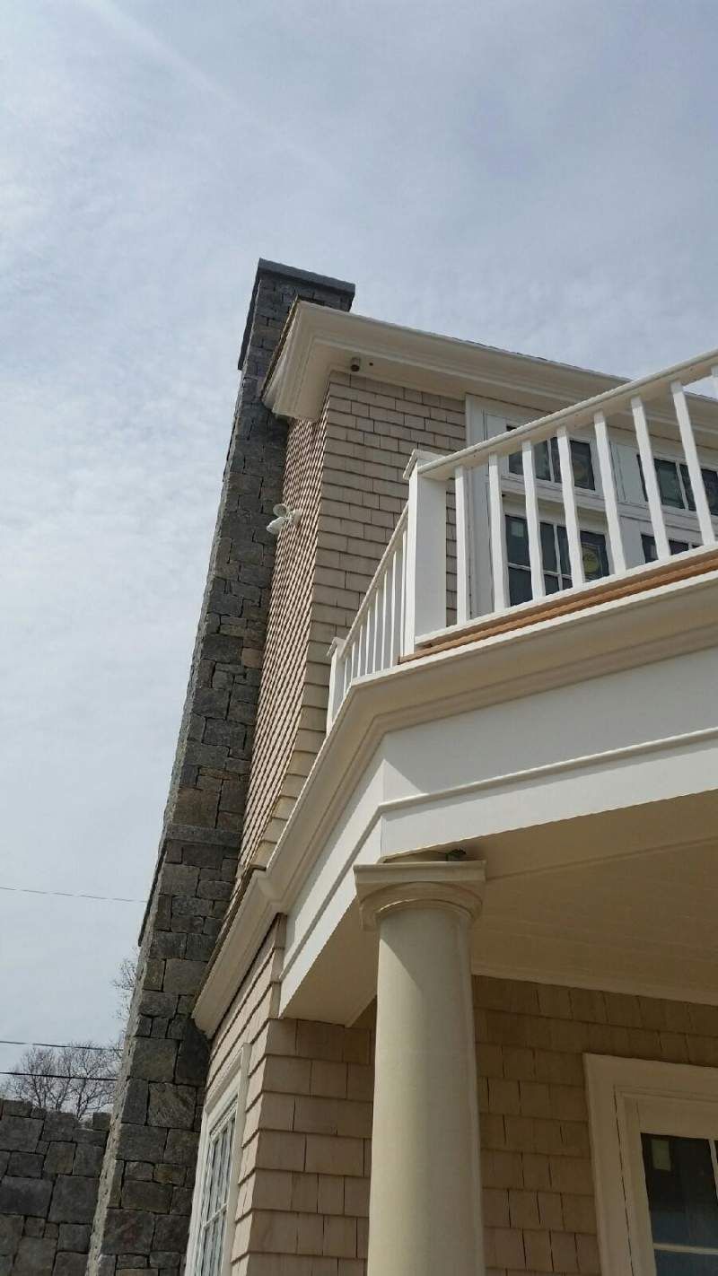 Stone chimney next to a beige house with a white balcony and column, under a cloudy sky.