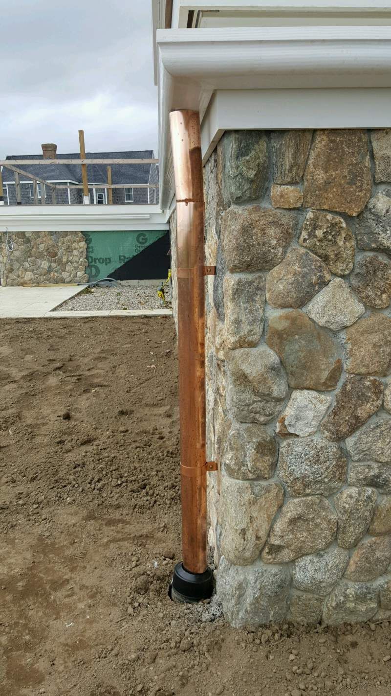 Copper downspout attached to a stone wall, extending to a black base on the ground.