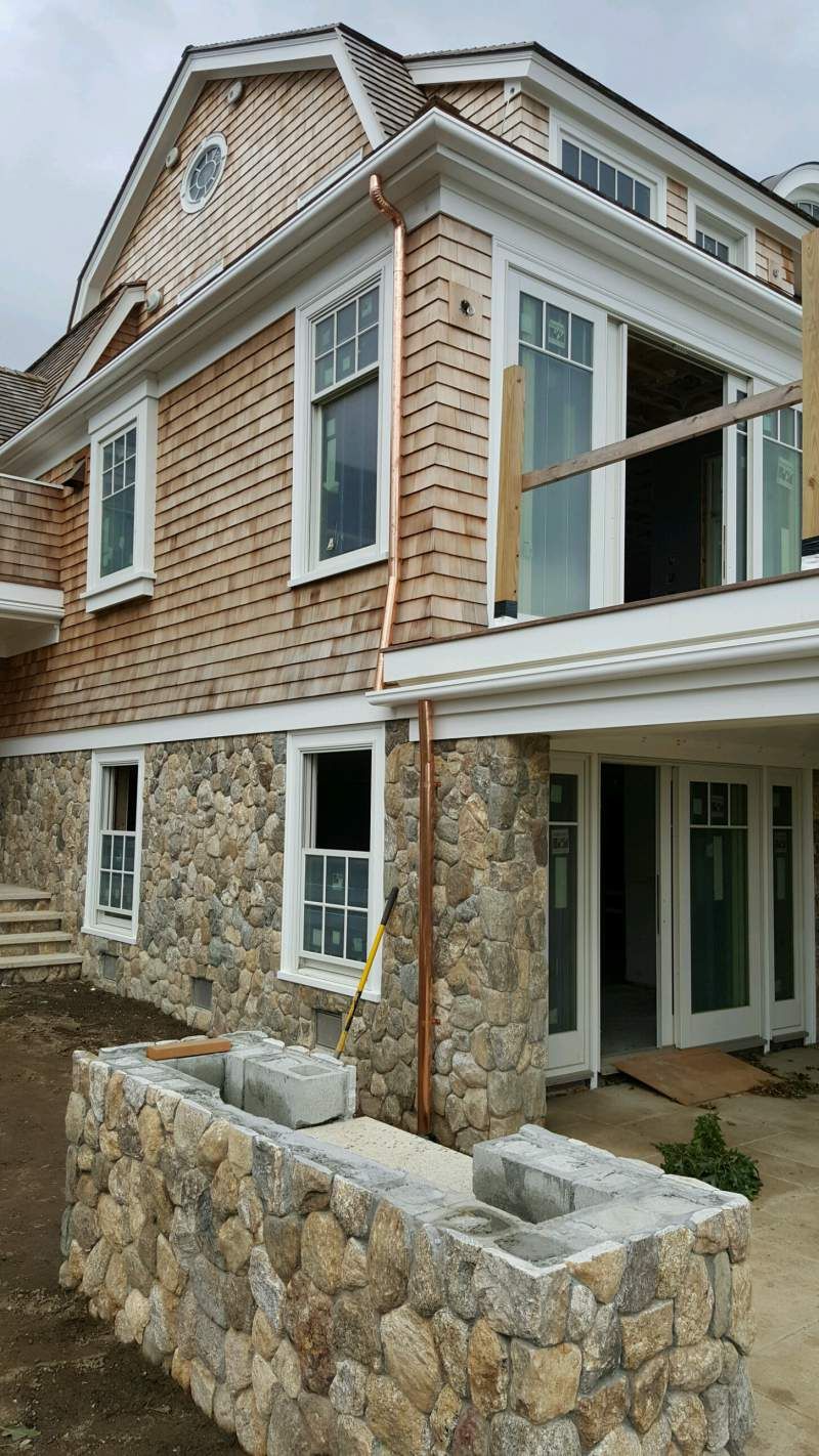 Two-story house with stone base and cedar shingle siding, white trim, and multiple windows; outdoor construction in progress.