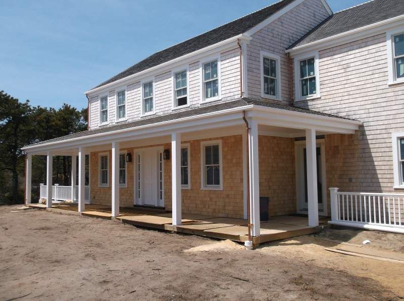 Two-story house with a large porch and light brown cedar siding on a sunny day.
