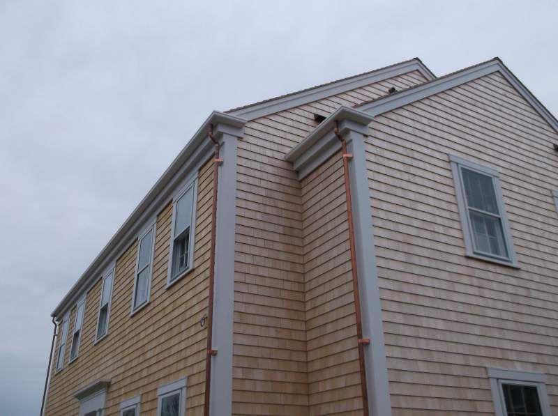 Two-story house with light brown shingle siding, white trim, and copper gutters against a cloudy sky.