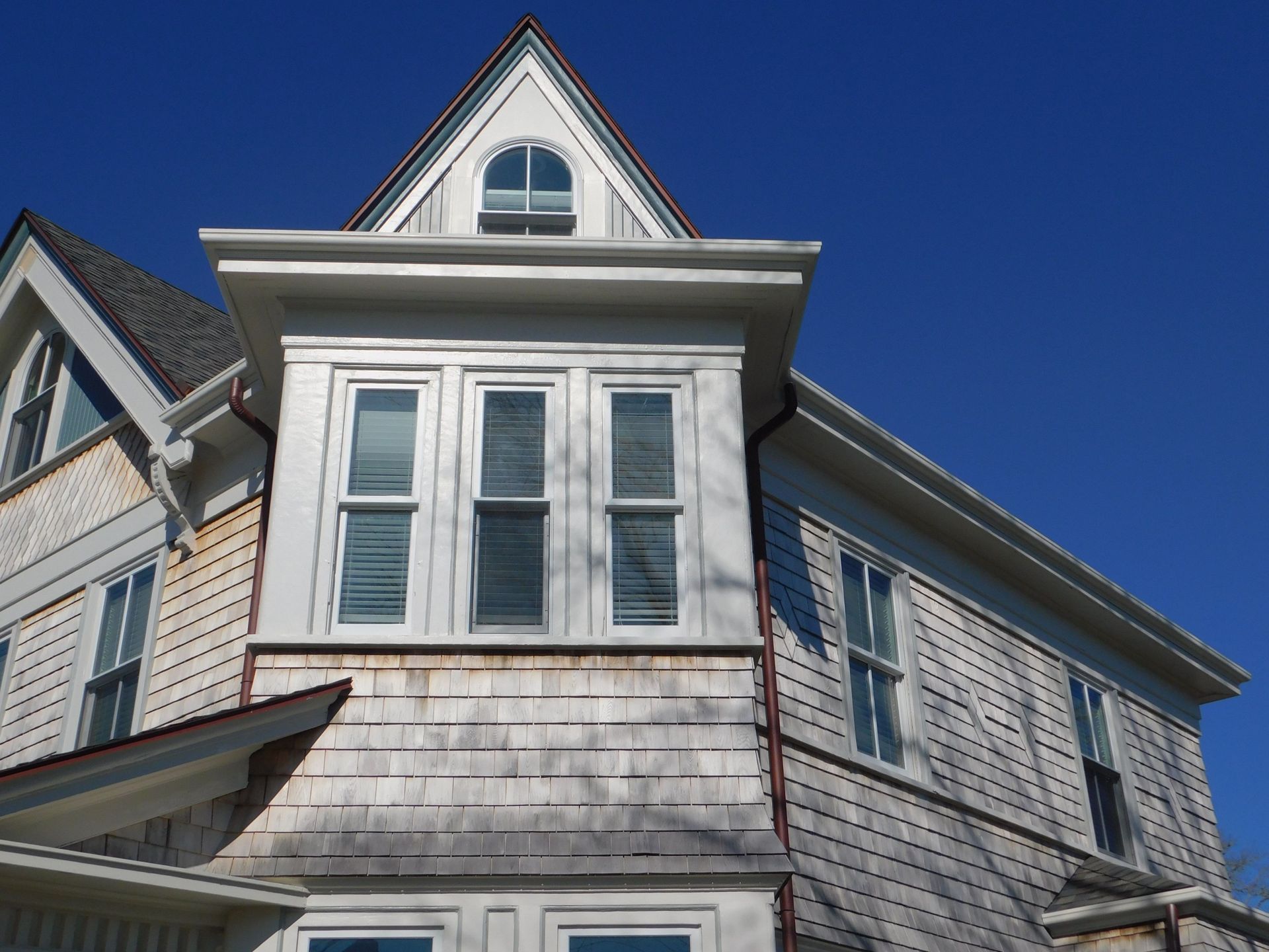 White shingled house exterior with a bay window and triangle-shaped top, against a blue sky.