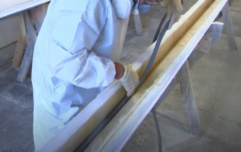 Person in protective gear using a saw to cut a long, light-colored object on a sawhorse in a workshop.
