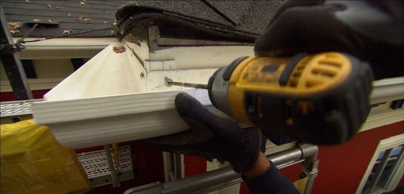 A person using a yellow drill to attach a white gutter to a house. They wear black gloves.