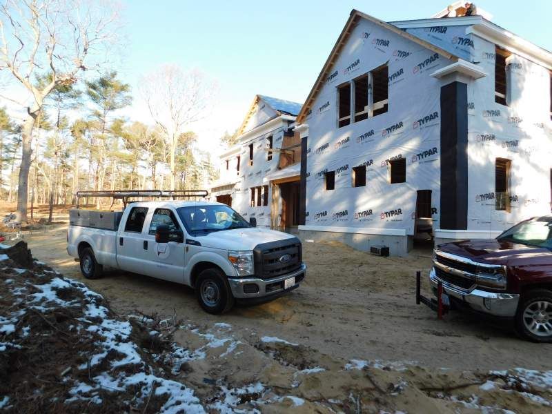 White Ford truck and red Chevy truck parked in front of a house under construction; snowy ground.