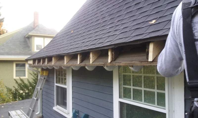 Construction worker repairing roof edge with exposed wood supports and dark shingles.