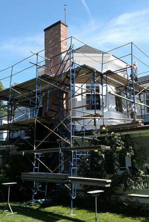Scaffolding surrounds a white house and brick chimney on a sunny day.