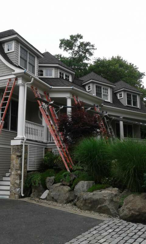 Three ladders leaning against a gray house's roof and porch, possibly for gutter work. Overcast day.