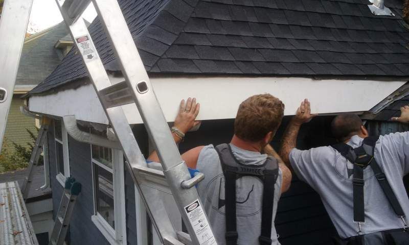 Two people installing white trim on a house roof overhang. A ladder is present.