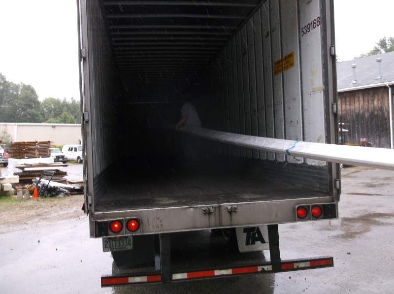 A person loads long, metal poles into a large, open cargo container; exterior of container and building visible.