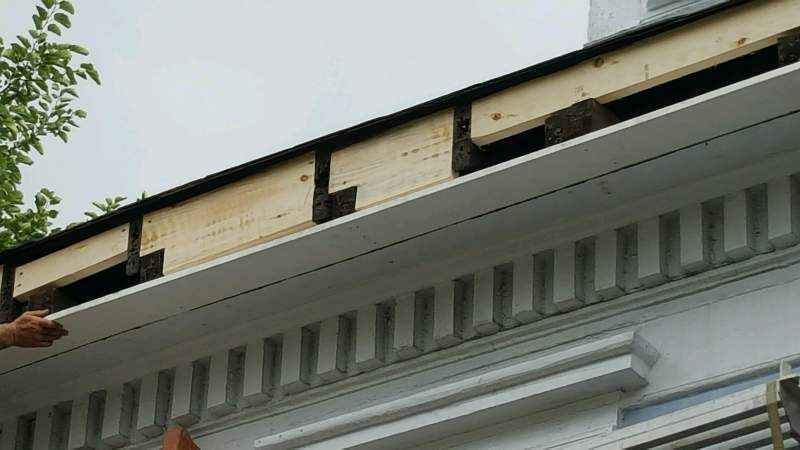 Installing a white soffit panel under a building's eave. Wooden framing is visible. Cloudy sky.