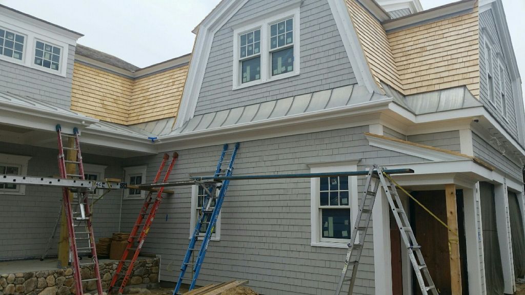 House under construction with gray siding, windows, and ladders. Beige roof partially installed. Cloudy day.