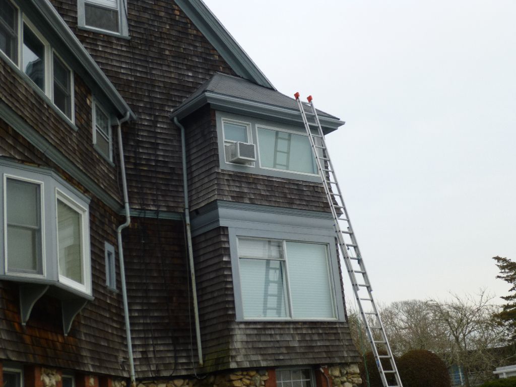 Ladder against a weathered shingled house. Windows with a/c units. Gray sky.