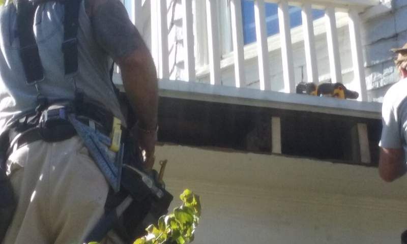 Workers repairing a house's exterior; one holds tools, another drills. White railing and siding visible.