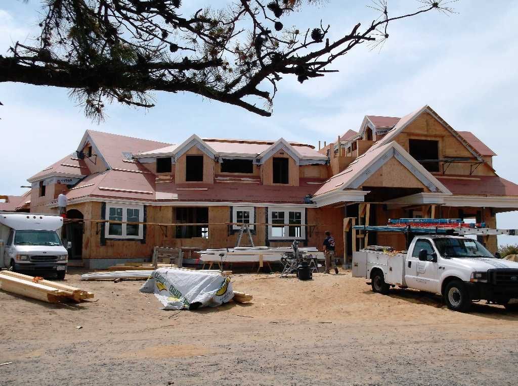 House under construction, wood framing and red roof, white work trucks parked outside.