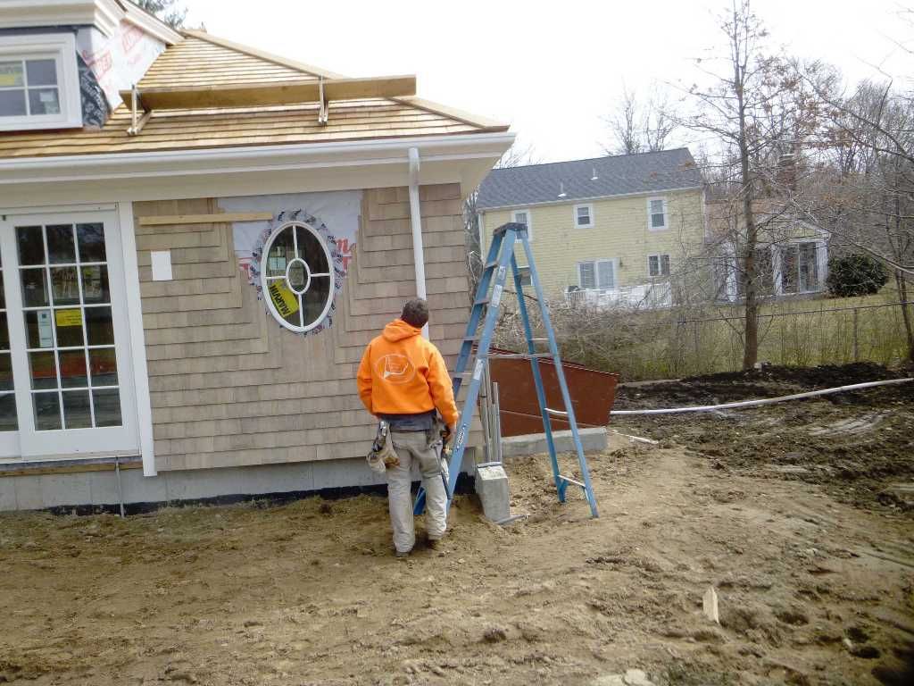 A construction worker in an orange jacket near a house with brown shingles and a blue ladder.