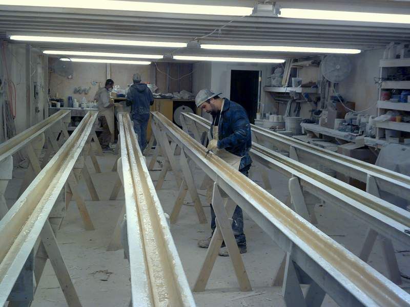 Workers in a workshop, applying a substance to long, trough-like structures supported by wooden frames.