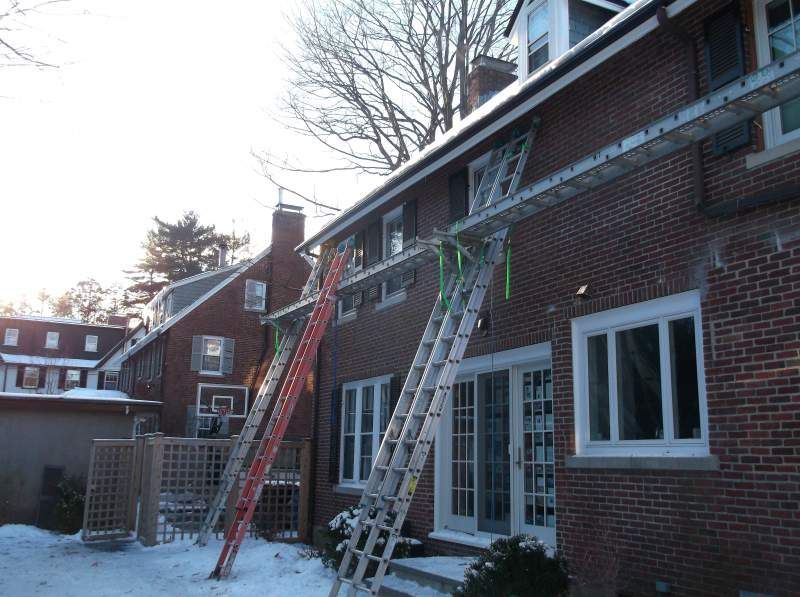 Ladders against brick buildings; snow on the ground. Two ladders reach roof and windows.