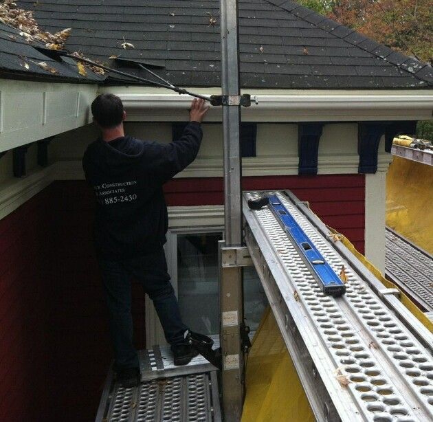 Man cleaning gutters with a tool while standing on a platform, red house in the background.