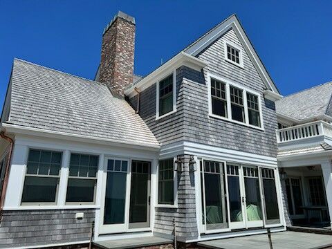 Gray shingled house with white trim, windows, and a brick chimney under a blue sky.