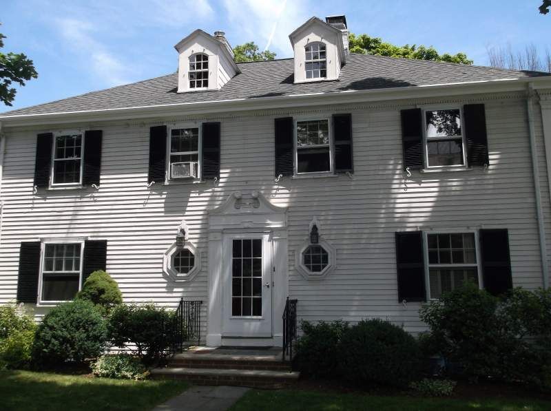 White two-story house with black shutters, dormers, and front door. Landscaping and bushes in front.
