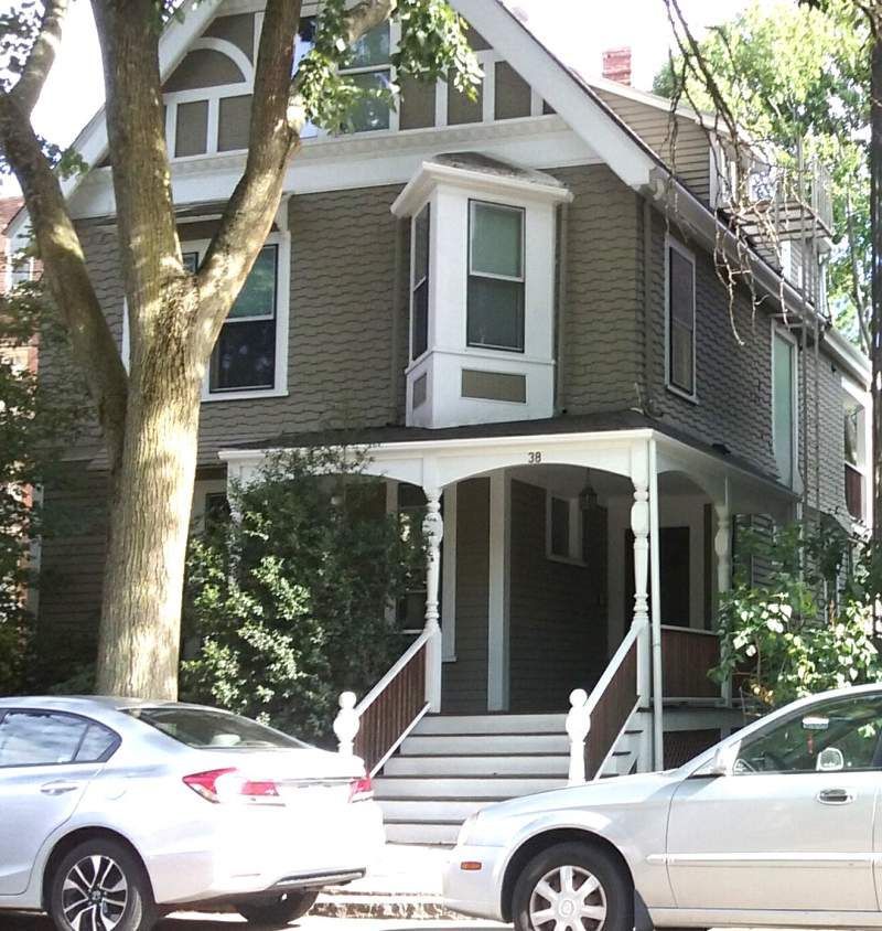 Two-story brown house with white trim, porch, and bay window; two cars parked in front, tree to the left.