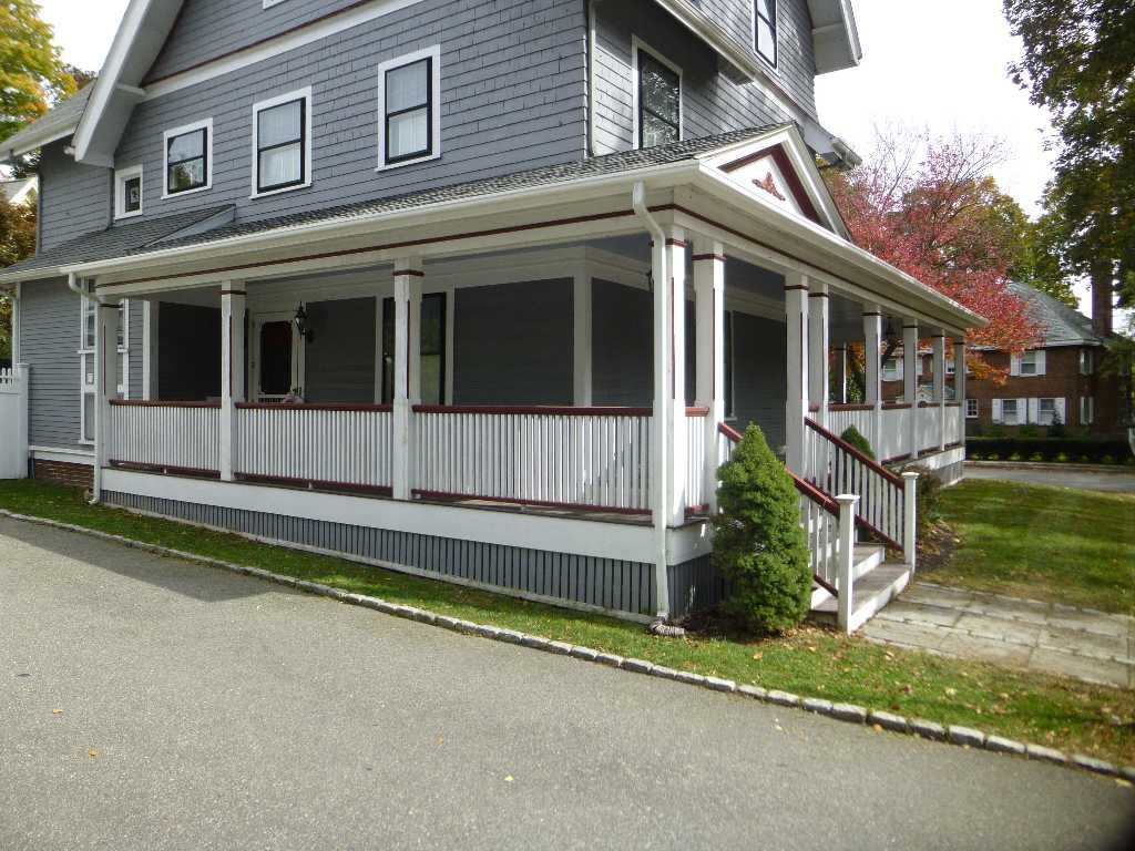 Gray house with a large porch. White trim, dark railings, and a small evergreen bush. Driveway in foreground.