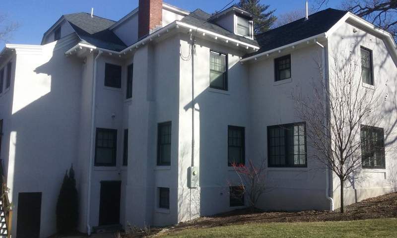 White stucco building with dark windows, chimney, and a small tree on a grassy hill.