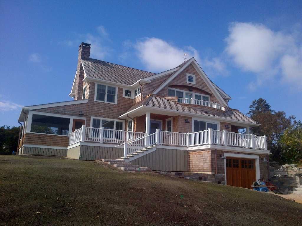 Two-story house with cedar shake siding, white railings, and a garage on a grassy hill under a blue sky.