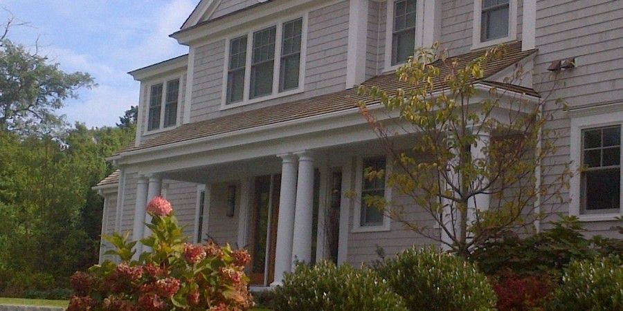 A gray shingled house with white columns and trim, bushes and a tree in front, under a blue sky.