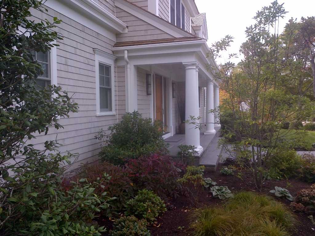 Beige house with a covered porch supported by white columns, surrounded by landscaping.